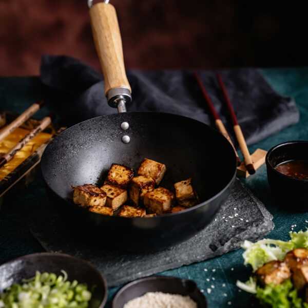 Gebratene Tofuwürfel im schwarzen Wok mit Sesam und Frühlingszwiebeln auf grünem Untergrund